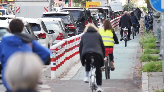 Einige Menschen fahren mit dem Fahrrad auf einem geschützten Radfahrstreifen. Daneben ist eine dicht befahrene Straße mit vielen Autos.