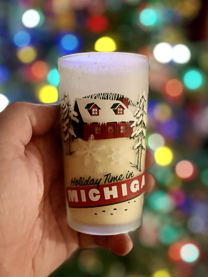 Photograph of a frosted glass of eggnog in a hand with a graphic image of a snowy cabin and “Holiday Time in MICHIGAN” written across the bottom. Christmas lights are visible in the background.
