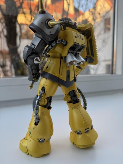 A detailed model of a yellow robot, viewed from the back, showcasing mechanical features, including pipes and a backpack-like attachment. The background features blurred buildings and trees.