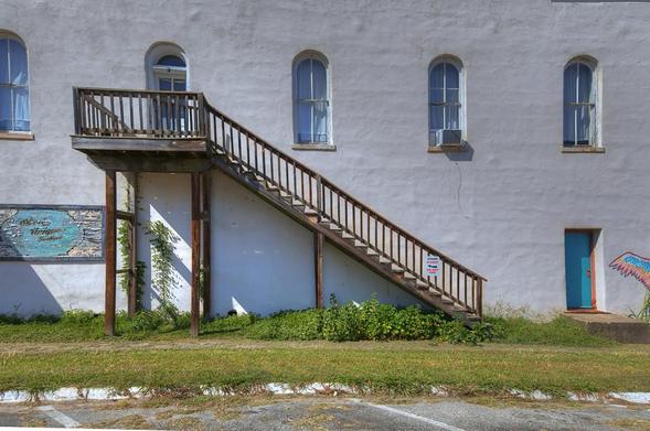 An old wooden staircase leans against a white building, with arched windows and a small door featuring colorful wings painted nearby.