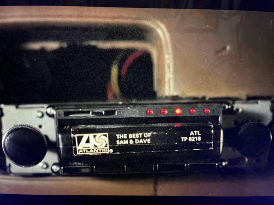 Image of a vintage tape player displaying an 8-track cassette titled "The Best of Sam & Dave" from Atlantic Records, featuring red indicator lights and a control knob on the left. The tape playing device sits uninstalled from a hole in the dashboard of a decommissioned cop car from mount prospect, circa 1978. 