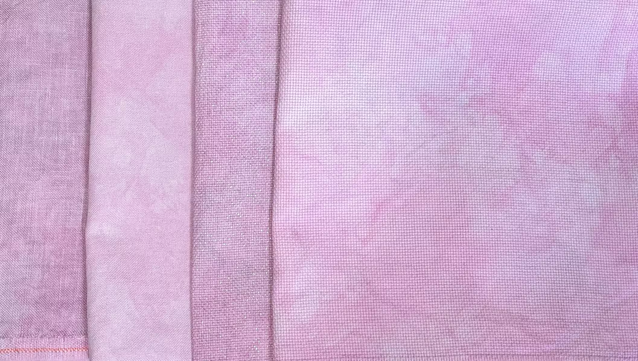 Four overlapping pieces of fabric in the same color (a dusty pink), but different types of fabric. Linen, evenweave, opalescent, and Aida.