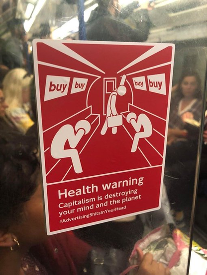 Sign on a London Underground train that has an outline sketch of a subway train with passengers shown bracing themselves from the advertisement banners above that have the words “buy, buy, buy, buy”.

Title: Health warning
Subtitle: Capitalism is destroying your mind and the planet