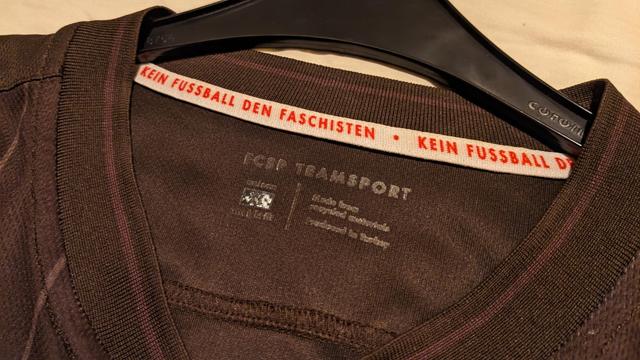 Bildbeschreibung von KI:
Das Bild zeigt ein dunkelbraunes T-Shirt mit dem Aufdruck „Kein Fussball den Faschisten“.

Das Bild zeigt einen Nahaufnahmeausschnitt eines dunkelbraunen T-Shirts, das auf einem schwarzen Kleiderbügel hängt.  Das Shirt ist aus einem feinmaschigen Stoff gefertigt.  Am Kragen des Shirts befindet sich ein weißes Etikett mit der Aufschrift „KEIN FUSSBALL DEN FACHISTEN“ in roter Schrift.  Die Schrift ist deutlich lesbar und wiederholt sich, wobei man den vollständigen Satz nur teilweise erkennen kann. Unter dem weißen Etikett ist ein kleinerer, dunkelgrauer Aufnäher mit der Aufschrift „FCSP TEAMSPORT“ sichtbar, sowie kleinere, kaum lesbare Textilkennzeichnungen.  Der Hintergrund ist unscharf und zeigt einen hellen,beigefarbenen Untergrund, möglicherweise ein Bettlaken oder eine Tischdecke.  Die allgemeine Atmosphäre ist neutral und sachlich, der Fokus liegt ganz auf dem Text auf dem T-Shirt, der eine klare politische Botschaft vermittelt. Die Stimmung ist eher ernst und politisch aufgeladen.