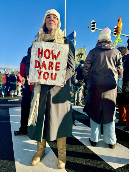 Vrouw staat op zebrapad met bordje in handen met tekst ‘how dare you’ erop. Blauwe winterlucht en veel andere demonstranten om haar heen.  