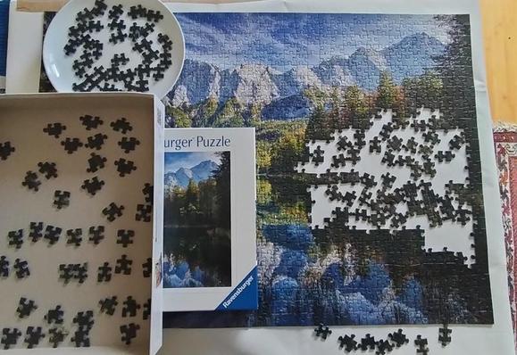 Puzzle vom Eibsee und dem Wettersteinmassiv mit der Zugspitze mit Lücke im rechten Bereich. Überall Puzzleteile auf einem Teller, in der Schachtel und in der unfertigen Fläche.