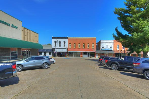 A small town street on the square scene features historic buildings and parked cars under a clear blue sky. 