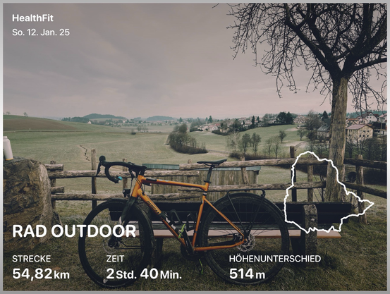 Im Vordergrund sieht man ein orangenes Fahrrad, das an einer Bank lehnt, im Hintergrund, Hügel,wälder und eine Ortschaft. Der Himmel ist bewölkt und es ist sehr kalt. Links und rechts des Bildes sieht man Grafiken über die Strecke die gefahrenen Kilometer und die Zeit.
