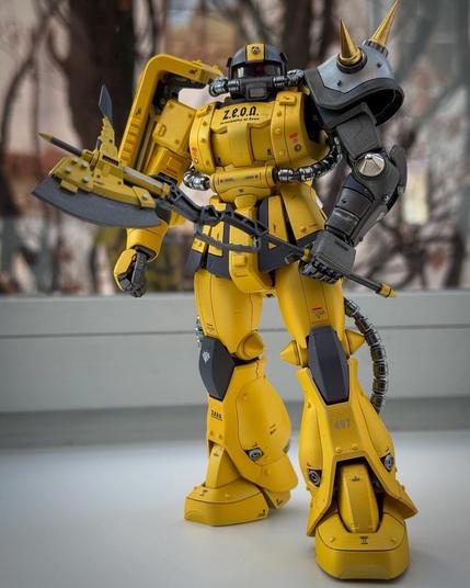 A detailed model of a yellow robotic figure, equipped with a large weapon and a spiked shoulder. The figure stands on a flat surface, with a blurred background of trees and buildings.