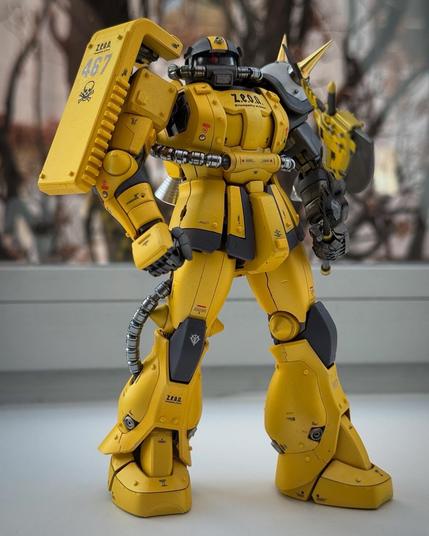 A yellow robotic model figure with a detailed design, including a shield and various mechanical elements. The figure stands on a white surface with a blurred background.