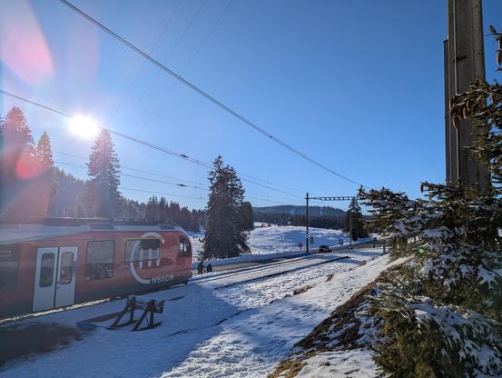 Un train NStCM à La Givrine prêt au départ pour La Cure. Paysage enneigé, grand soleil, ciel bleu, contrastant avec le rouge du train.