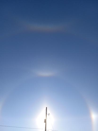 Looking up at the sky, with nested rainbow arcs around the sun, which is blocked by a telephone pole.  