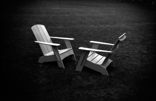A black and white photograph of two Adirondack chairs facing each other on a field of grass.