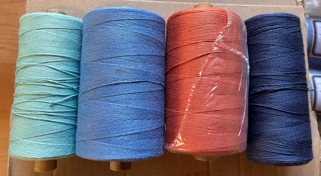 4 cones of 8/2 cotton weaving yarn in robin’s egg blue, slate blue, rust, and navy sitting on a weaving bench