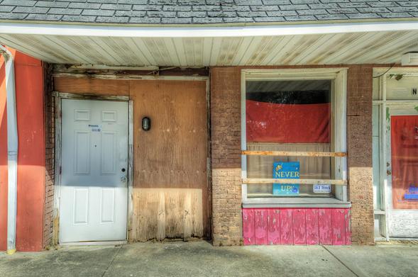 An old storefront displays a faded red curtain and a motivational sign in the window.