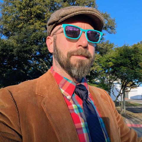 A photo of me, a 46 year old white man with a shaved head, a short cropped brown & grey beard, 

photographed from the chest up in front of some live oak trees in an office park,

wearing

a rectangle glasses with turquoise frames, a red, turquoise and navy blue plaid dress shirt, a navy bkue necktie in a trinity knot under a tan corduroy blazer, with a light brown flat cap