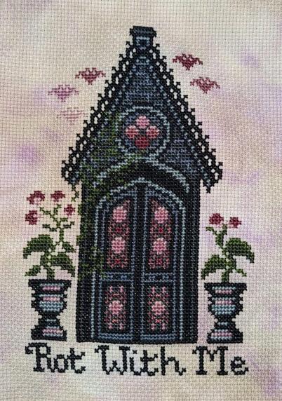 A finished cross stitch of a small mausoleum with pink stained glass. Two larger planters with pink flowers are on either side of the door and pink bats fly overhead. Below the mausoleum are the words "Rot With Me"
