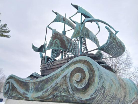 It appears to be a two masted ship in bronze, but now all that faded greenish color. It sits on a swirly motif that probably represents ocean. 

The ship is in full sail, and there are the netting kinds of ladder things that go up to each mast. 