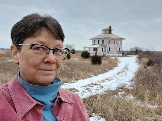 I'm a brown haired white woman wearing glasses, with a slight smile to the camera for the selfie. Behind my left shoulder is the Pink House, a decrepit two story squarish house with no windows left. It does appear to have a light blue door intact on one side.

A snow covered dirt drive curves between me and the house. Nearby long marsh grasses and some shrubs sit around the house. 

I am wearing a pink shirt with a blue turtleneck underneath. I did not know beforehand that I would match the house.  