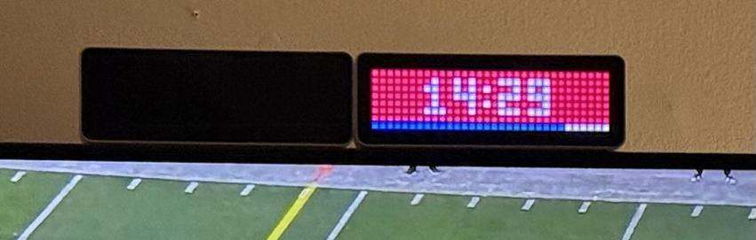An LED matrix display showing a countdown timer and progress bar sitting on top of a tv showing an (unrelated) football game. The display is in the colours of the home team. 