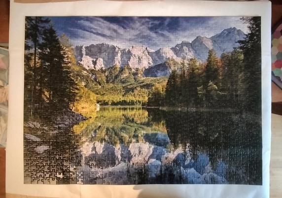Puzzle mit 1000 Teilen zeigt den Eibsee mit dem Wettersteingebirge und der Zugspitze. Eingerahmt von Herbstwald liegt der glatte See da. Im Hintergrund ragen die schroffen Felswände aus weißem Kalkstein in die Höhe, darüber ein blauer Himmel mit weißen Federwolken. Der Wald am Seeufer im Vordergrund ist sehr dunkel. Berge, Wolken und Wald spiegeln sich in der Mitte des Bildes perfekt im See. 