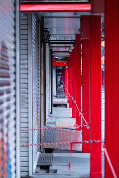 A perspective shot down the covered shopping arcade of Nakamise-dori at Sensoji Temple in Tokyo on a rainy morning. The traditional walkway features bright red support columns on one side and closed metal shop shutters on the other. The empty corridor creates a dramatic vanishing point, with red and white caution tape crossing the space. A solitary figure with an umbrella can be seen in the distance, capturing the quiet, early morning atmosphere before the usually bustling shopping street opens for the day.