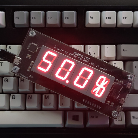 **TL;DR:** A digital "Short Life" clock displaying "50.0%" over a mechanical keyboard, reminding the user that half of their expected life is gone.

The clock features a red LED display reading "50.0%" with a glitch-like graphic in the final digit. Above the display is the text: *"SHORT LIFE — A clock to remind you that life is short."* The device is placed on a white mechanical keyboard with a visible Windows key and function keys (F5-F11).