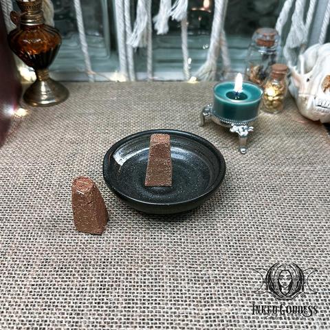 An example of the "Ceramic Dish with Large Cone Incense" product are placed on a burlap-covered surface. It features a small glazed ceramic dish and two large incense cones. The dish is dark grey and the cones are brown and shaped like pyramids with flat tops. Presented by Inked Goddess Creations.