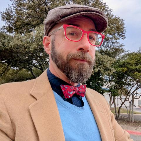A photo of me, a 46 year old white man with a shaved head, a short cropped brown & grey beard, 

photographed from the chest up in front of some live oak trees in an office park,

wearing

rectangle glasses with red frames, a red bowtie with light blue paisleys on a navy blue dress shirt under a sky blue v-neck sweater under a camelhair blazer, with a light brown flat cap
