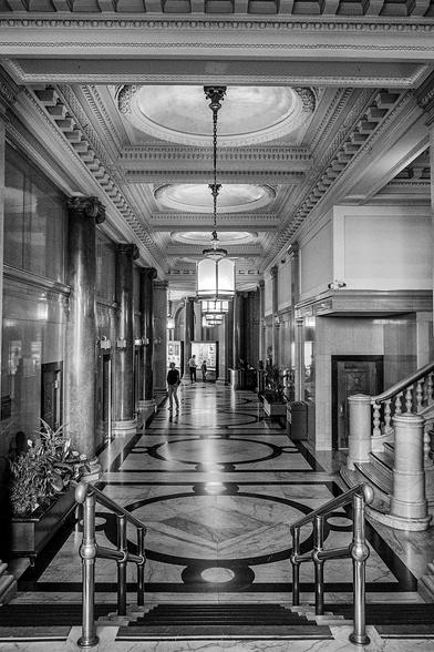 Ornate lobby with chandeliers, marble floors and marble columns. A bank of elevators is to the left and a set of stairs is to the right. 