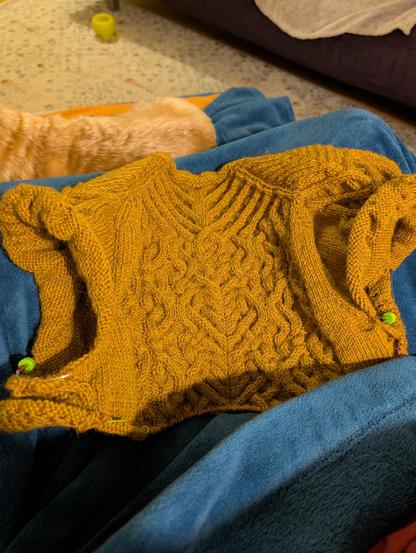 View from the top of a knitting project using mustard yellow yarn and featuring a lot of cables in its design. It's setup on a dark teal blanket on my lap.