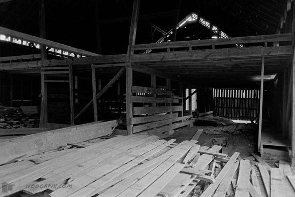Black and white photograph of the interior of a barn, with scattered wooden planks on the floor and a loft area above. The light filters through gaps in the wooden slats, creating a pattern of shadows.