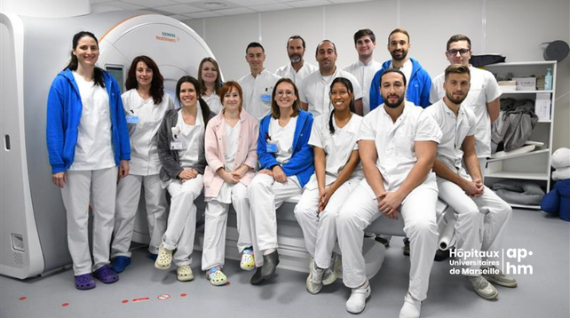 Photo de l'équipe IRM de l'Hôpital de la Timone (AP-HM), une partie du service d'imagerie médicale, composée de 14 professionnels de santé en tenue blanche, posant devant un appareil IRM.