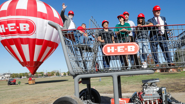 Groundbreaking ceremony featuring H-E-B Grocery Store executives in red H-E-B hard hats standing in a giant shopping cart and waving. In the background, a red & white striped H-E-B hot-air balloon looms gracefully.