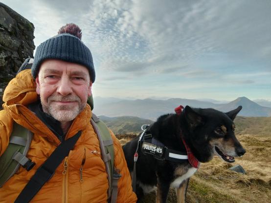 Selfie at the summit trig point, Moray the dog and me. I'm wearing a woollen hat and warm clothing against the wind. In the distance more hills. Hazy cloudy sky.