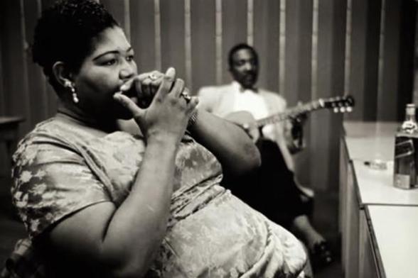 Big Mama Thorton, tocando la armónica en un camerino. Es una mujer afroamericana voluminosa que lleva un vestido ceñido a flores y tiene anillos en las manos. Al fondo, desenfocado, un guitarrista también afroamericano con pajarita y chaqueta descansa con una guitarra en las manos. A la derecha, sobre una mesa, destaca una botella de whisky o bourbon. Dominio público.