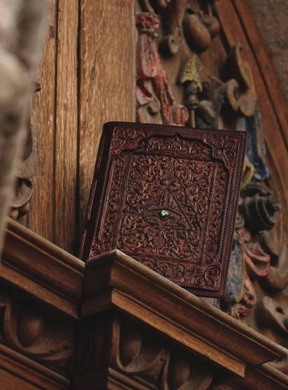 Photo featuring the front cover of leather notebook covers. Covers are decorated by all sorts of floral and curved carvings and there is additional layer of leather frame to make it even more impressive and massive. There is an eye-like triangular symbol at the center which in turn has a green precious stone installed in it. Color of the covers is mahogany. Photo background is colorful carved medieval door sill.