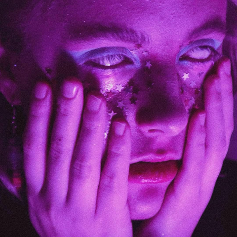 portrait d’une personne qui a un maquillage avec des paillettes étoile et qui se tient le visage, les yeux totalement blancs