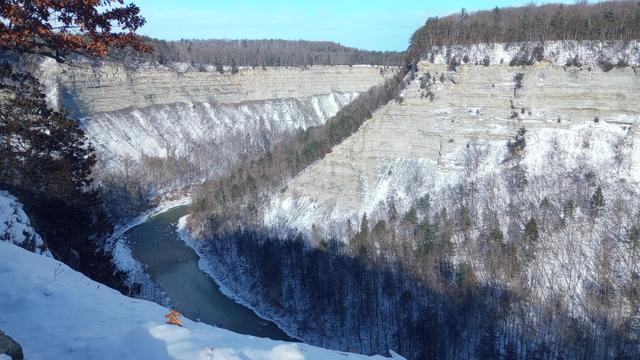 A deep gorge with an icy river cutting through below. Steep rock faces are occasionally broken by clusters of snow capped trees.