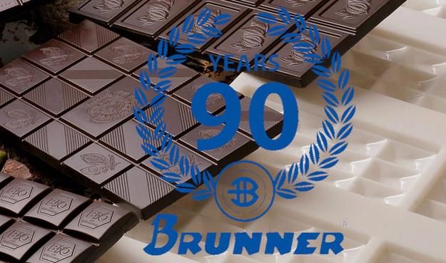 Zentral ein Blauer Kranz mit der Schrift 90 Jahre darunter der Name Brunner. Im Hintergrund verschiedene dunkle Schokoladentafeln und leere weiße Schokoladenformen.