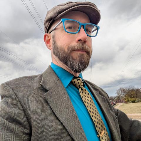 A photo of me, a 46 year old white man with a shaved head, a short cropped brown & grey beard, 

photographed from the chest up in front of some live oak trees in an office park,

wearing

a rectangle glasses with teal frames, a gold necktie with black diamonds in a trinity knot on a teal dress shirt under an olive herringbone blazer and a light brown flat cap