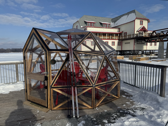 Geo some of wood and plastic containing 4 red lawn chairs. A large building in the background at the edge of a frozen river