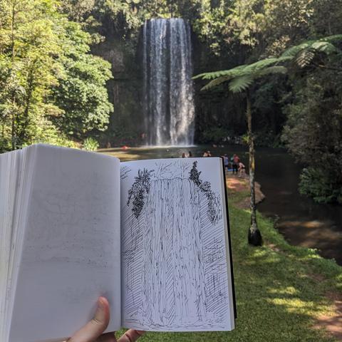 A photograph of a tropical waterfall in the background, with a sketch of that waterfall in a sketchbook held in the foreground