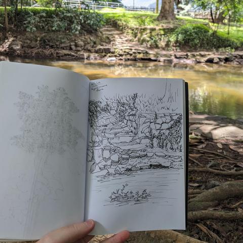 A photograph of stone steps leading down to a creek in the background, with a sketch of the steps and creek in a sketchbook held in the foreground