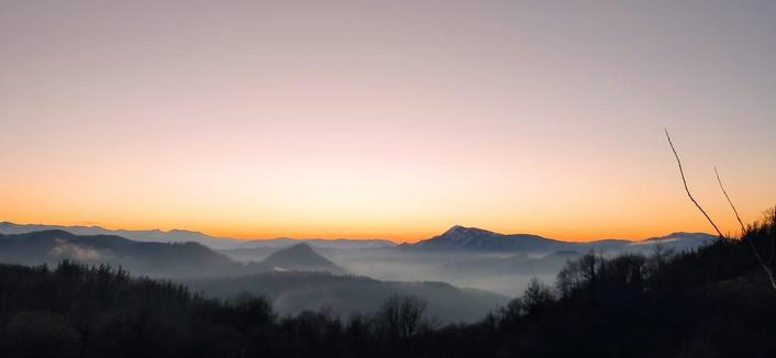 
Una puesta de sol anaranjada y dorada se extiende sobre una cadena montañosa silueteada.  Las montañas se extienden hasta el horizonte, con una capa de neblina o niebla baja en los valles.  En primer plano, se aprecian árboles y arbustos oscuros contra el cielo brillante.  Una rama seca destaca en la esquina derecha. La escena evoca una sensación de calma y tranquilidad.