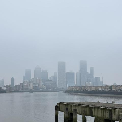 A photo of the Canary Wharf skyline taken from further down the Thames path. We're looking east, and Canary Wharf's high-rise buildings are shrouded in fog. In the foreground, an old, abandoned pier structure struts out into the river.