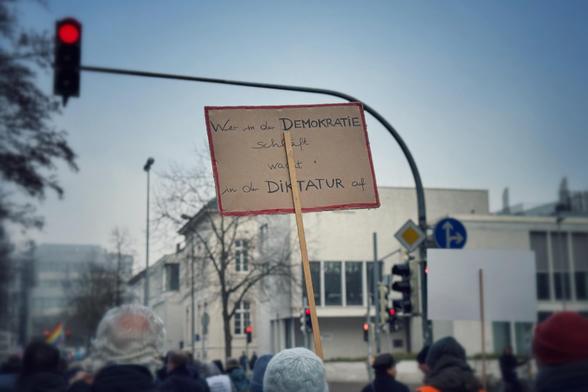 Während einer Demo auf einer Hauptstraße mit roter Ampel im Hintergrund hält jemand ein Pappschild mit dem Schriftzug „Wer in der Demokratie schläft, wacht in der Diktatur auf“ in die Höhe. 