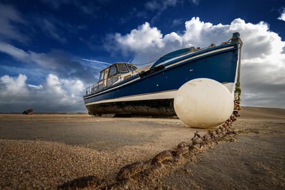 Die Fotografie zeigt ein trocken gefallenes Boot, das auf einem sandigen Strand liegt. Im Vordergrund ist eine große, weiße Boje zu sehen, die mit einer Kette mit dem Boot verbunden ist. Das Boot ist blau mit weißen Akzenten und hat ein kleines Steuerhaus. Der Himmel im Hintergrund ist blau mit einigen weißen Wolken. Die Perspektive des Fotos ist von unten aufgenommen, was dem Bild eine dramatische Wirkung verleiht.