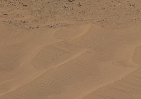 A rust toned photography of a Martian dune field. On close inspection, one spots a little helicopter stranded near the crest of one of the dunes.