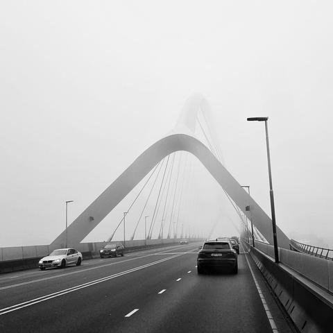 Zwart wit foto van een mistige achtergrond, half ovaal-rond poortachtige constructie boven een vierbaansweg autoweg met twee doorgetrokken strepen in het middenvanaf de weg genomen, lantarenpalen en weerzijden, enkele rijdende auto's aan weerzijden